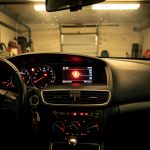 Tableau de bord de voiture dans une ambiance de garage, avec le voyant moteur allumé et des reflets sur le verre sous une lumière chaude.