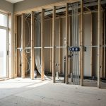 Intérieur d’une maison en rénovation complète, avec murs ouverts laissant apparaître les gaines électriques, la plomberie et la structure en bois, éclairé par la lumière naturelle.