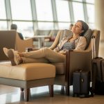 Une voyageuse assise dans un salon lumineux après un long vol, les jambes légèrement surélevées sur un repose-pieds, avec un bagage à ses côtés.
